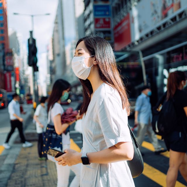 young asian woman with protective face mask using smartphone while commuting in downtown city street against crowd of pedestrians and city buildings