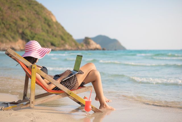 mujer con traje de baño y sombrero trabajando con un portátil en la playa