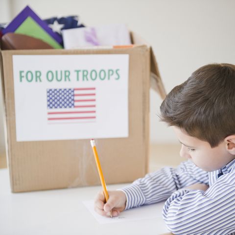 a little boy writing a letter with a pencil next to a donation box that says "for our troops" with a picture of the american flag