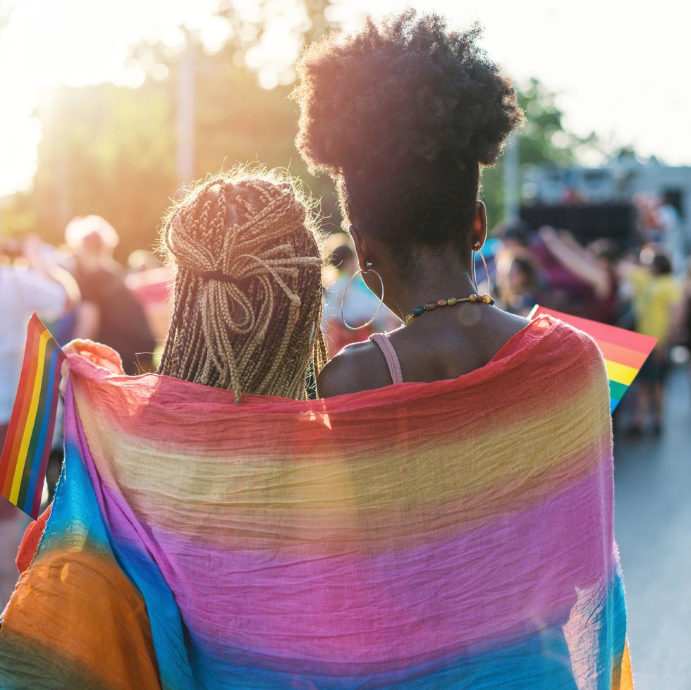 The picture focuses on two black women standing together in a side hug. They are both turned a way from the camera, and face a crowd of people. A big LGBTQ+ flag is draped around both of them, and each one holds a smaller flag. 