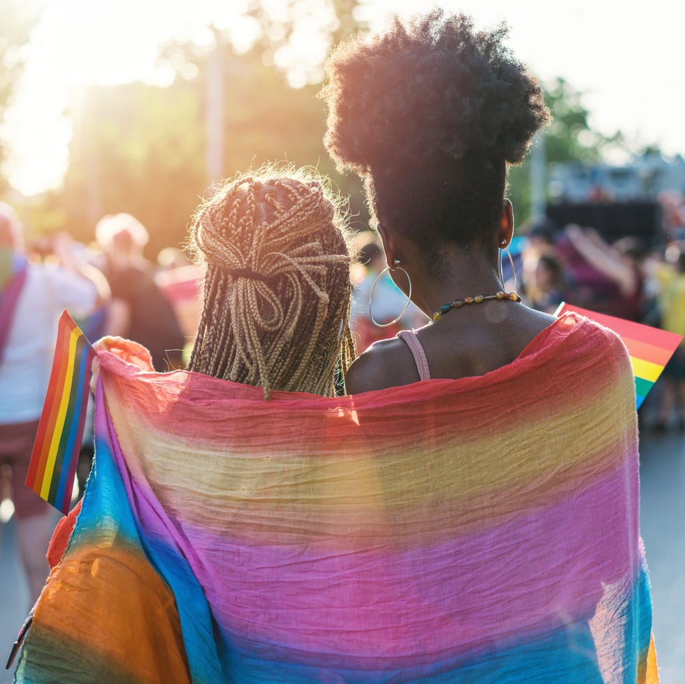 The picture focuses on two black women standing together in a side hug. They are both turned a way from the camera, and face a crowd of people. A big LGBTQ+ flag is draped around both of them, and each one holds a smaller flag. 