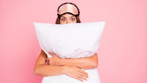 Young gorgeous shocked lady wearing eye mask, hugging pillow, hiding. Isolated over pink pastel background
