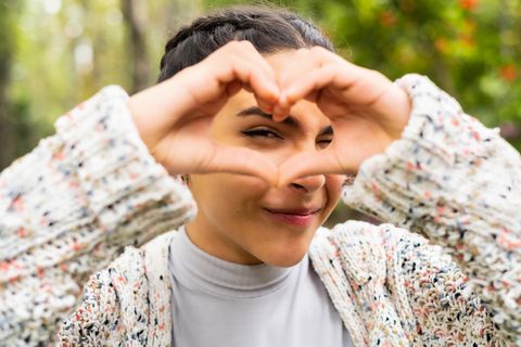 young latino woman smiling, looking at the camera and making a heart gesture with her hands, love