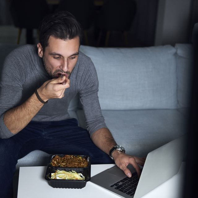 Young man sitting on sofa in evening eating takeaway and using laptop