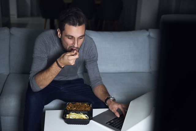 young man sitting on sofa in evening eating takeaway and using laptop