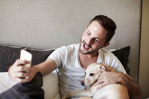 Young man taking smartphone selfie with dog on bed