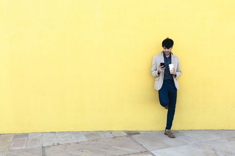 Young man with coffee to go checking the cell phone in front of yellow wall