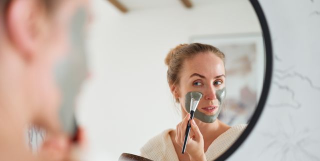 young woman applying clay beauty mask at her face standing against mirror in bathroom