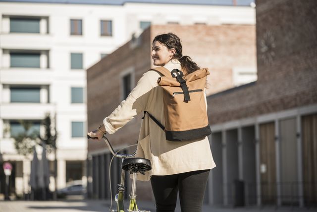 young woman carrying backpack while walking with bicycle on city street