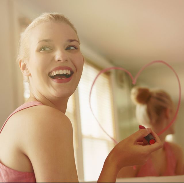 young woman in bathroom drawing heart shape on mirror with lipstick