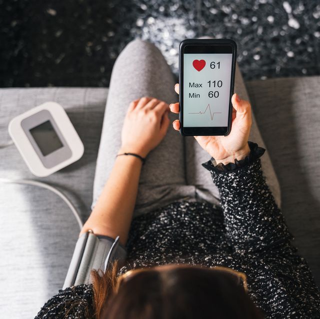 young woman measures blood pressure sitting on sofa at home with smartphone connected to device   concept of health, well being and love for oneself   millennial in a moment of private life