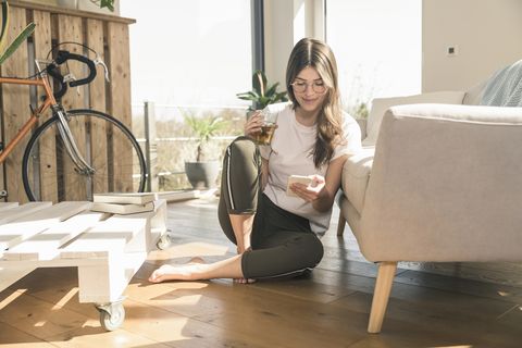 young woman sitting on the floor at home with drink and cell phone