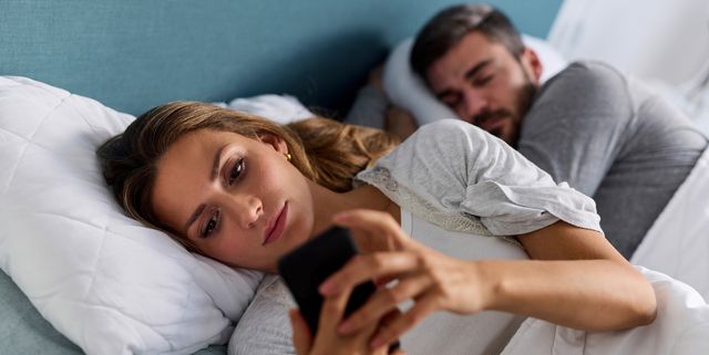 young woman using mobile phone and checking messages in bed while her husband is asleep