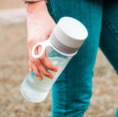 A young woman wearing a casual clothing is holding a reusable water bottle container while outdoor