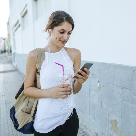 Young woman with cell phone and smoothie walking down the street