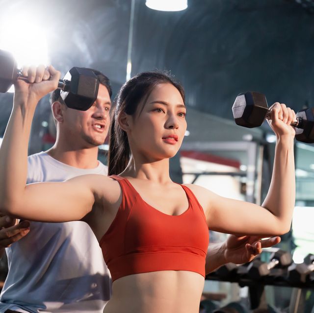 young woman with dumbbells and personal trainer flexing muscles in gym, sport and fitness concepts