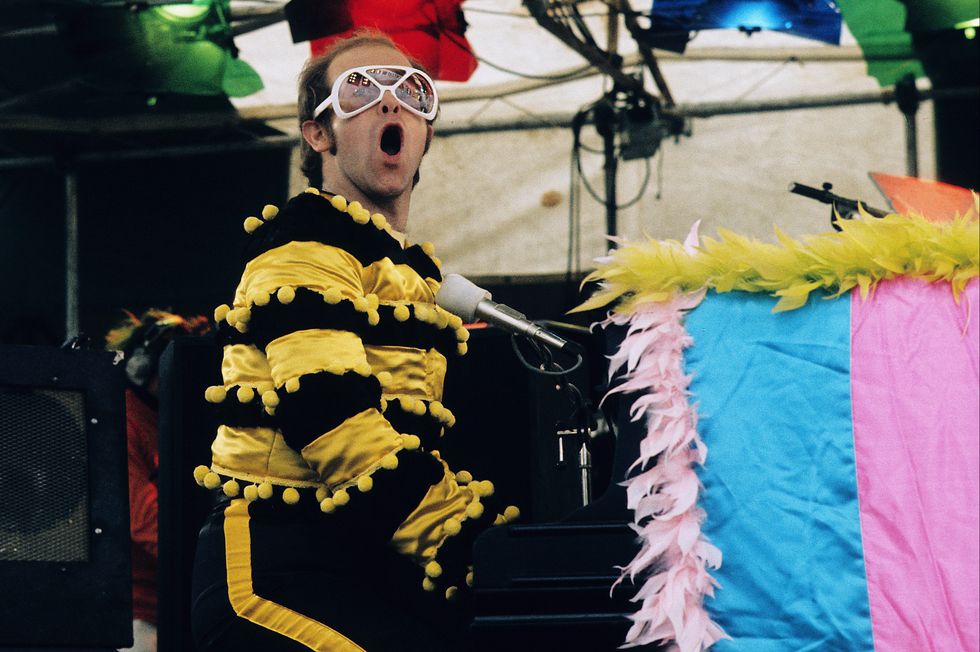 elton john sings while sitting and playing a piano, he wears a bee costume with glasses
