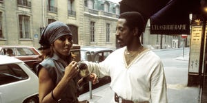 ike and tina turner stand on a sidewalk, tina is wearing a gray dress and bandana and looks at the camera, ike wears an all white outfit with a belted tunic and adjusts something on tinas dress