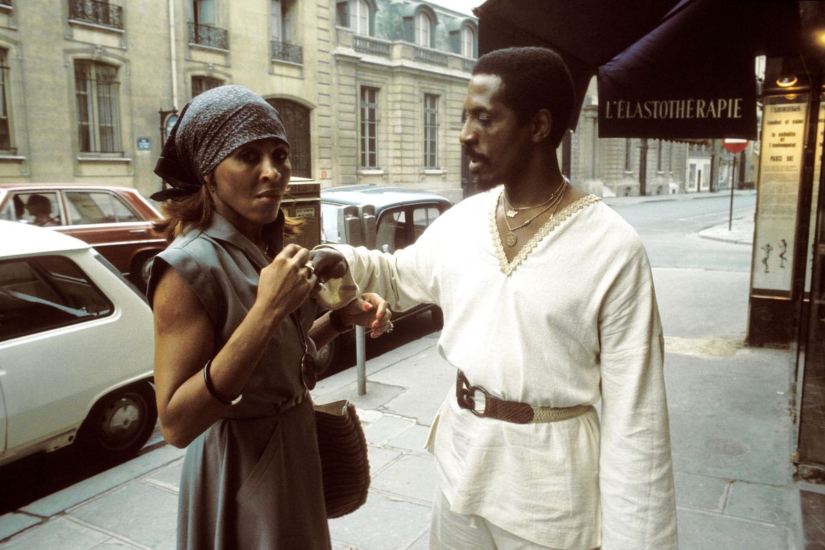 ike and tina turner stand on a sidewalk, tina is wearing a gray dress and bandana and looks at the camera, ike wears an all white outfit with a belted tunic and adjusts something on tinas dress