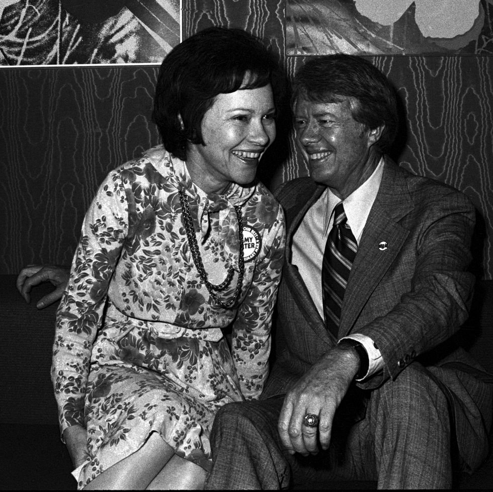rosalynn carter and jimmy carter sit on a couch and smile widely, she is wearing a floral dress with a campaign button, and he has on a suit