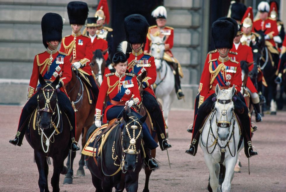 In 1984 neemt Elizabeth II te paard de parade Trooping the Colour af waarbij ze wordt vergezeld van haar zoon kroonprins Charles rechts van haar en wordt gevolgd door haar man en anderen Alle deelnemers zijn gekleed in uniformen van de Welsh Guards en gedecoreerd met koninklijke medailles De mannen dragen de hoge militaire berenmuts