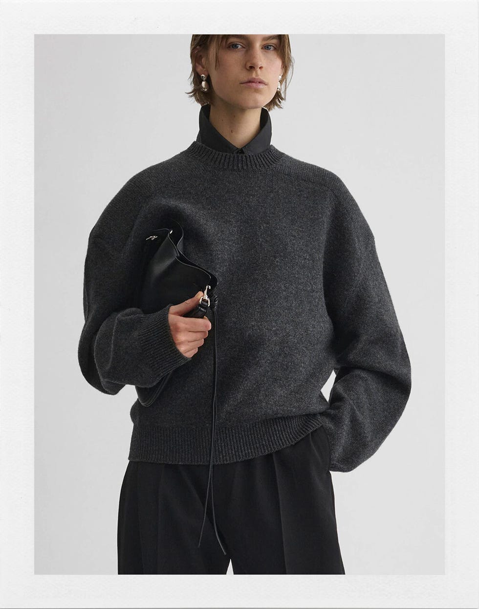 a model presents a dark gray sweater paired with black trousers holding a black clutch