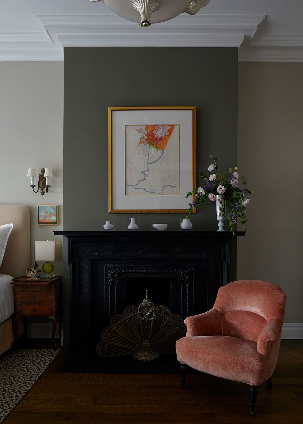 black mantel in bedroom with small white objects on it with a plush salmon chair in front and a piece of artwork above the mantel