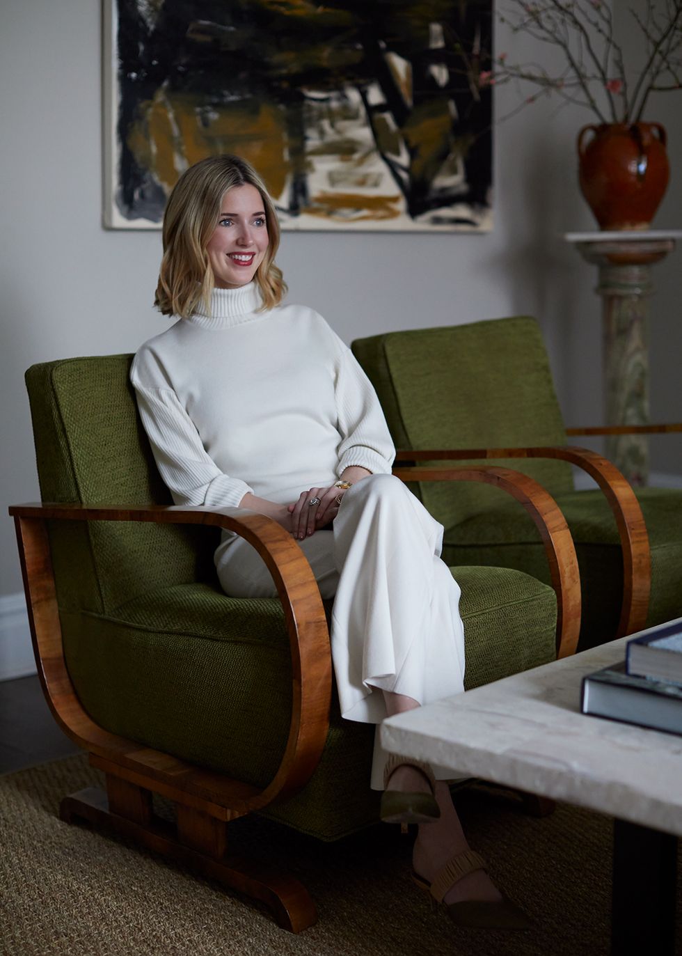 smiling designer waring all white turtleneck and flair pants sitting in a green armchair