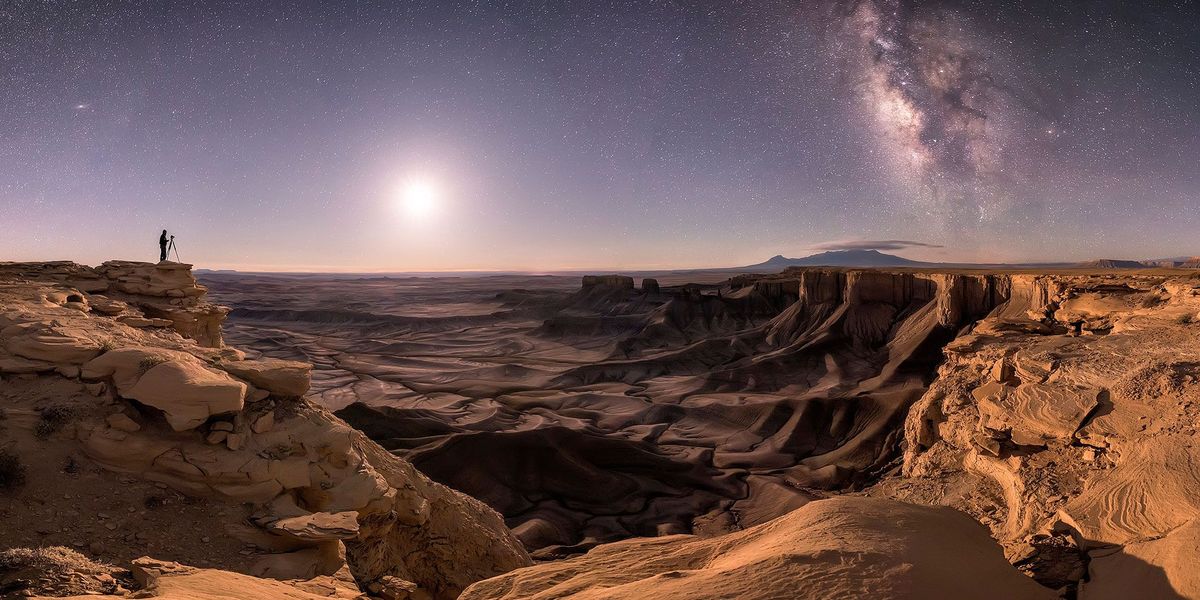 In Moab Utah steekt het silhouet van een eenzame fotograaf af tegen de nachthemel waar onder andere de maan en de sterrenstelsels Andromeda en de Melkweg zijn te zien