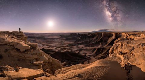 In Moab Utah steekt het silhouet van een eenzame fotograaf af tegen de nachthemel waar onder andere de maan en de sterrenstelsels Andromeda en de Melkweg zijn te zien