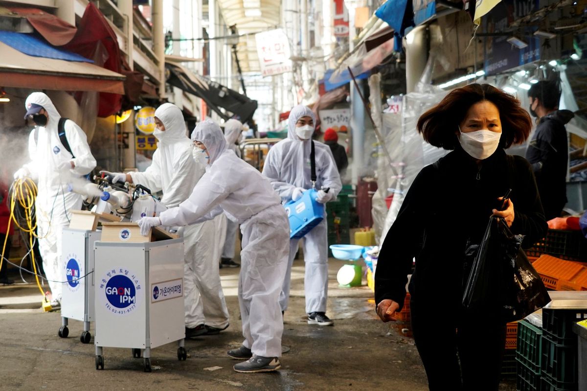 Een vrouw die een mondmasker draagt om besmetting met het nieuwe coronavirus te voorkomen loopt weg terwijl werknemers van een ontsmettingsbedrijf een traditionele markt in Seoul ZuidKorea desinfecteren 26 februari 2020