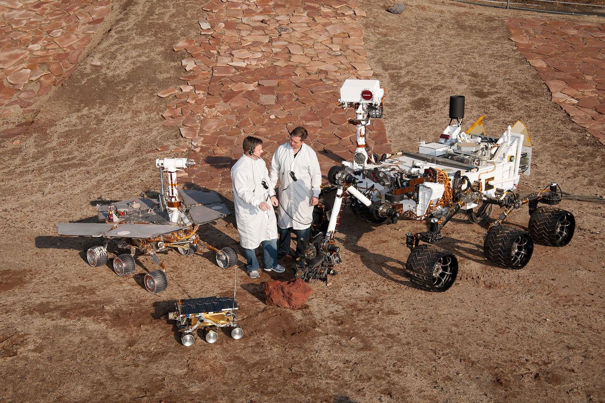 Twee NASAingenieurs poseren met drie generaties Marsrovers die ontwikkeld zijn in het Jet Propulsion Laboratory in Pasadena Californi De Curiosity rover was bedacht als een laboratorium op wieltjes zegt Vasavada