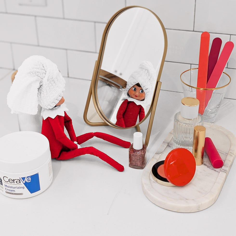 an elf on the shelf has a washcloth wrapped around its hair like a towel, sits in front of a dollhouse mirror and has cerave, nail polish and makeup spread around it