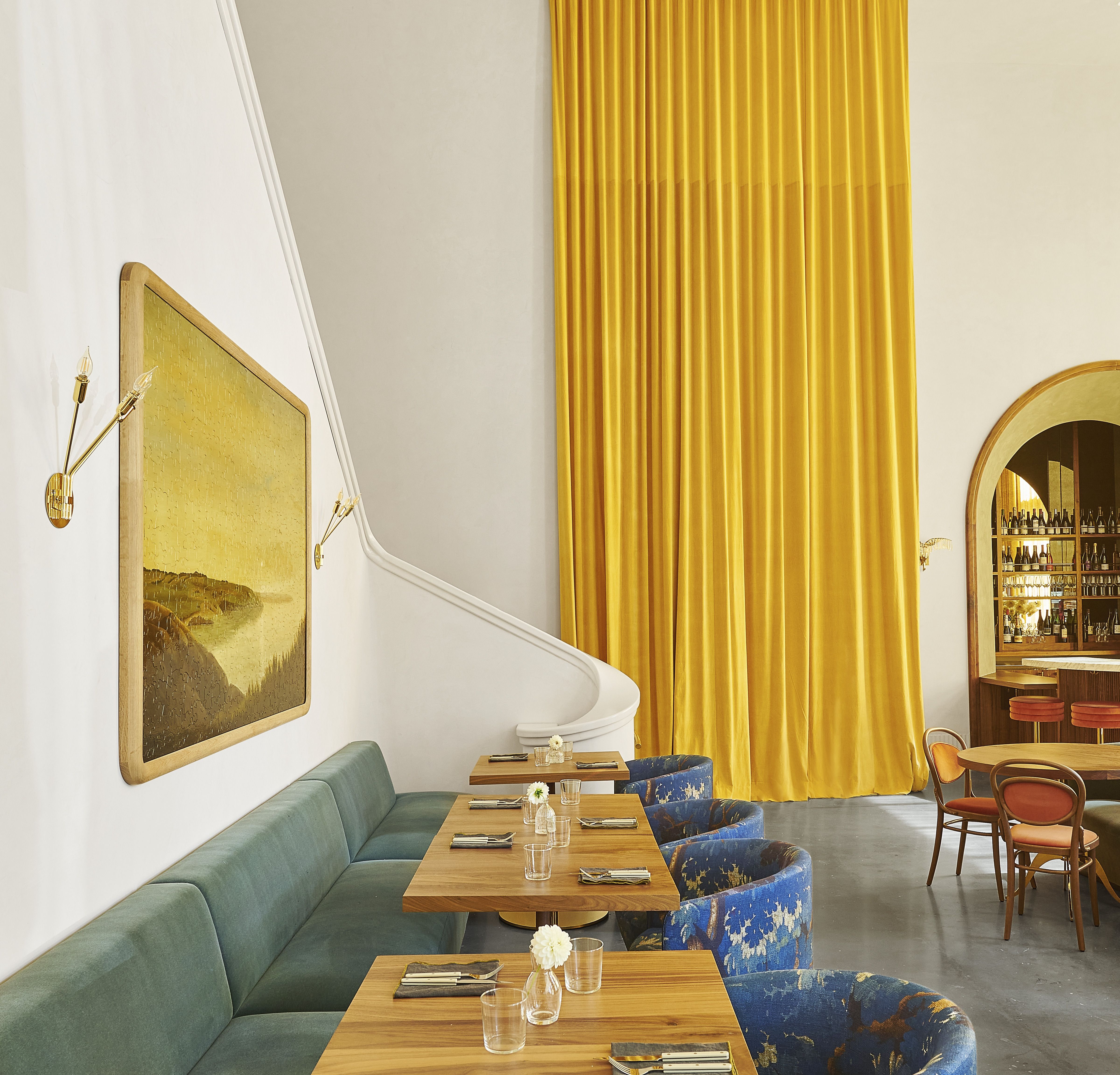 A List 2022 - ELLE DECOR interior of restaurant with green banquette long tables and round back chairs in blue with a long yellow curtain at the back and some orange chairs to the rifht
