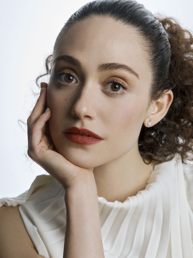 actress emmy rossum poses in a white top with her chin in her hand and her hair in a simple ponytail