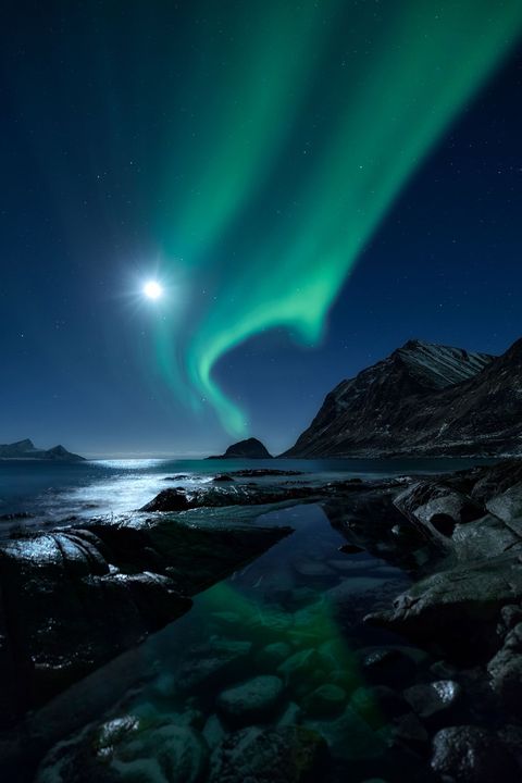 Ondanks het felle schijnsel van een extra heldere maan glanst het noorderlicht boven een fjord van de Noorse Lofoteneilanden