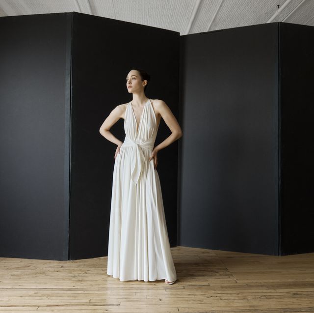 actress emmy rossum stands in a bare room in front of a black screen wearing a white grecian goddess gown