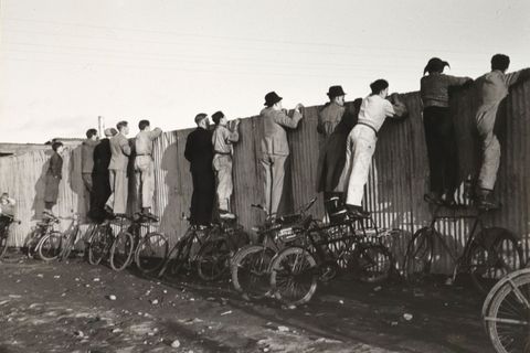 Balancerend op hun fietsen kijken IJslandse fans in 1943 over een schutting om een voetbalwedstrijd te volgen