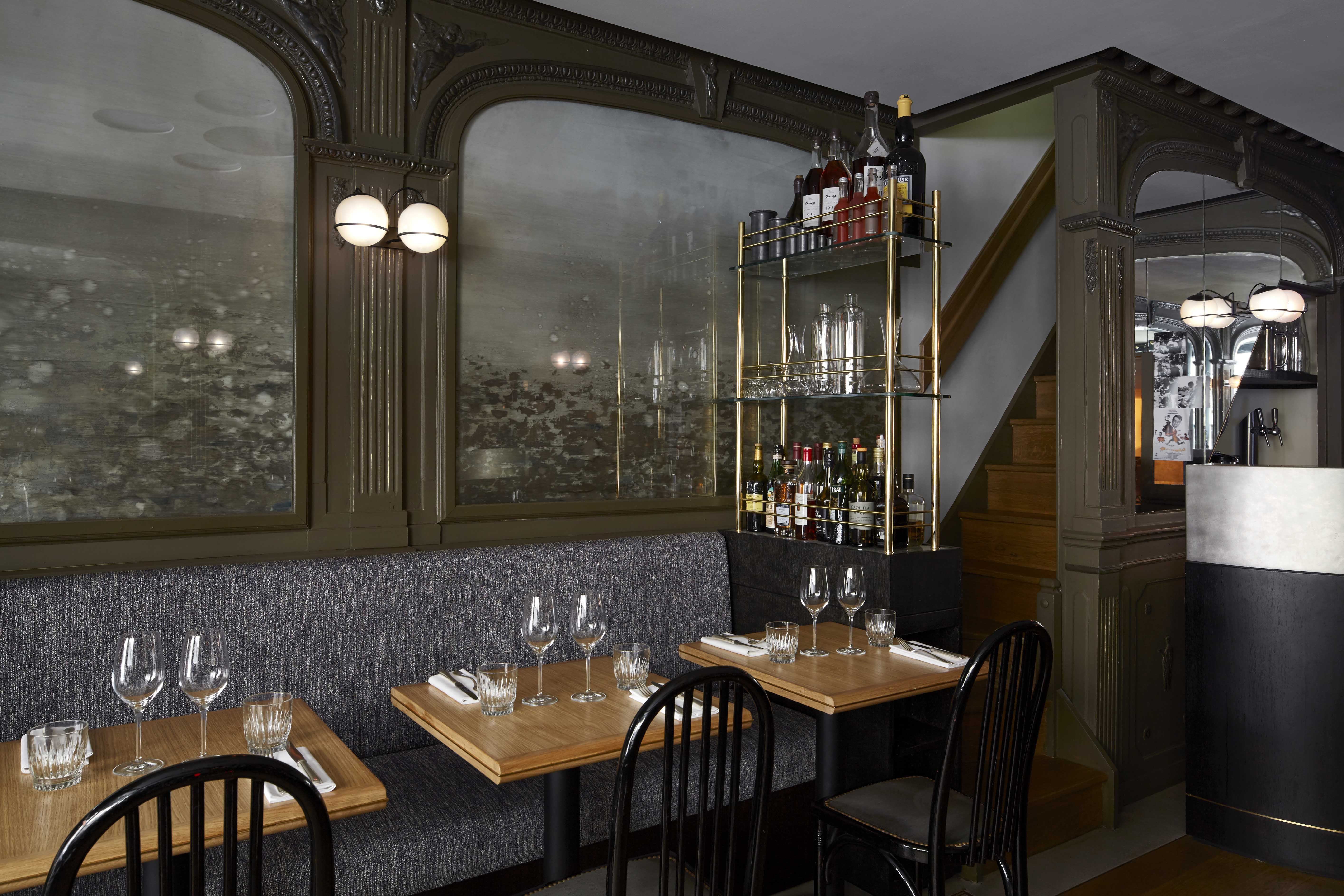 A List 2022 - ELLE DECOR dark interior of restaurant with small tables of two and a gray fabric banquette by the mirrored wall