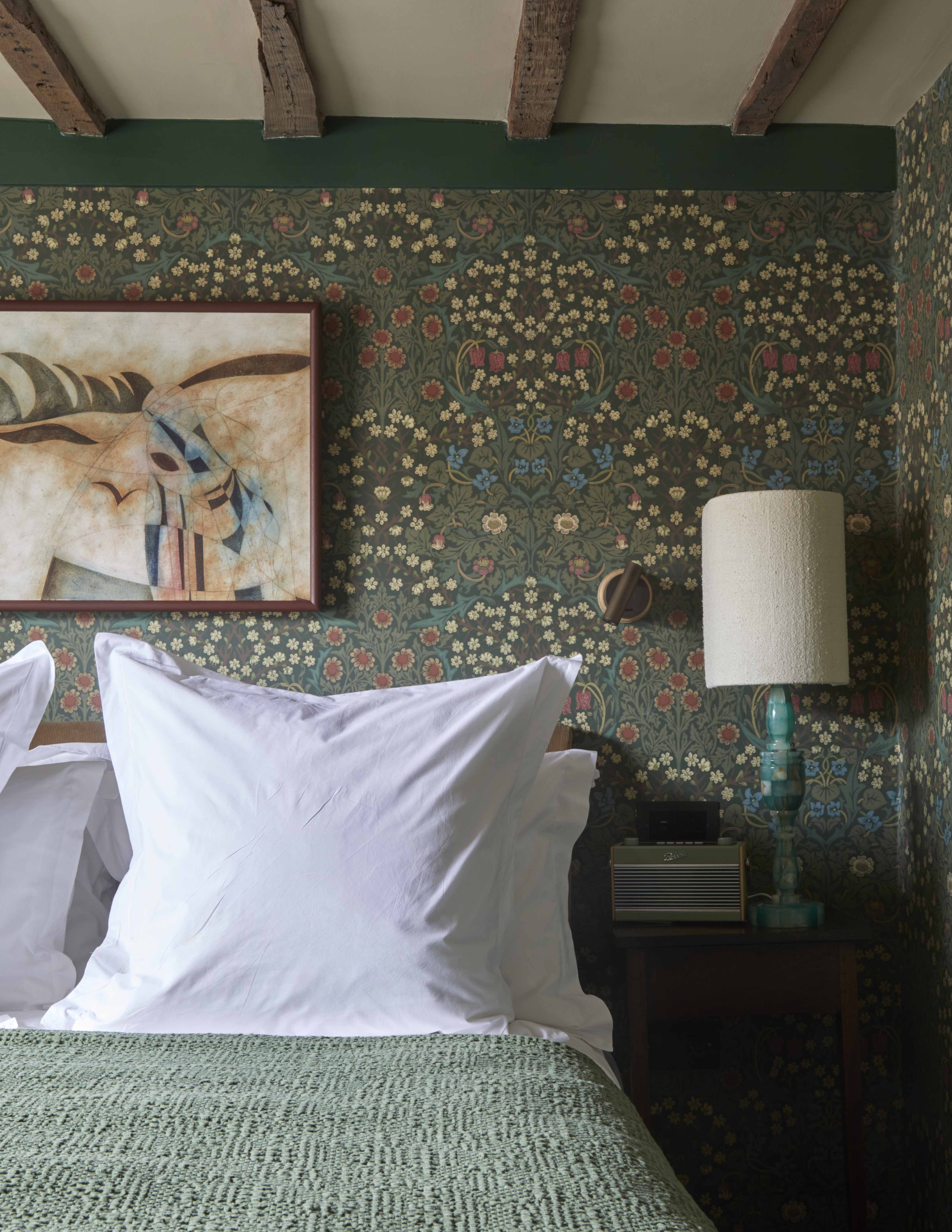 A List 2022 - ELLE DECOR bed with green coverlet and a flowery green wallpaper behind it and a nightstand with a blue lamp and artwork over the bed