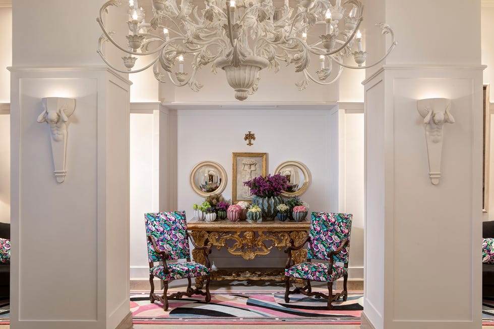 hotel interiors with two brightly upholstered armchairs, a white rococo chandelier, and a gilded table topped with plants