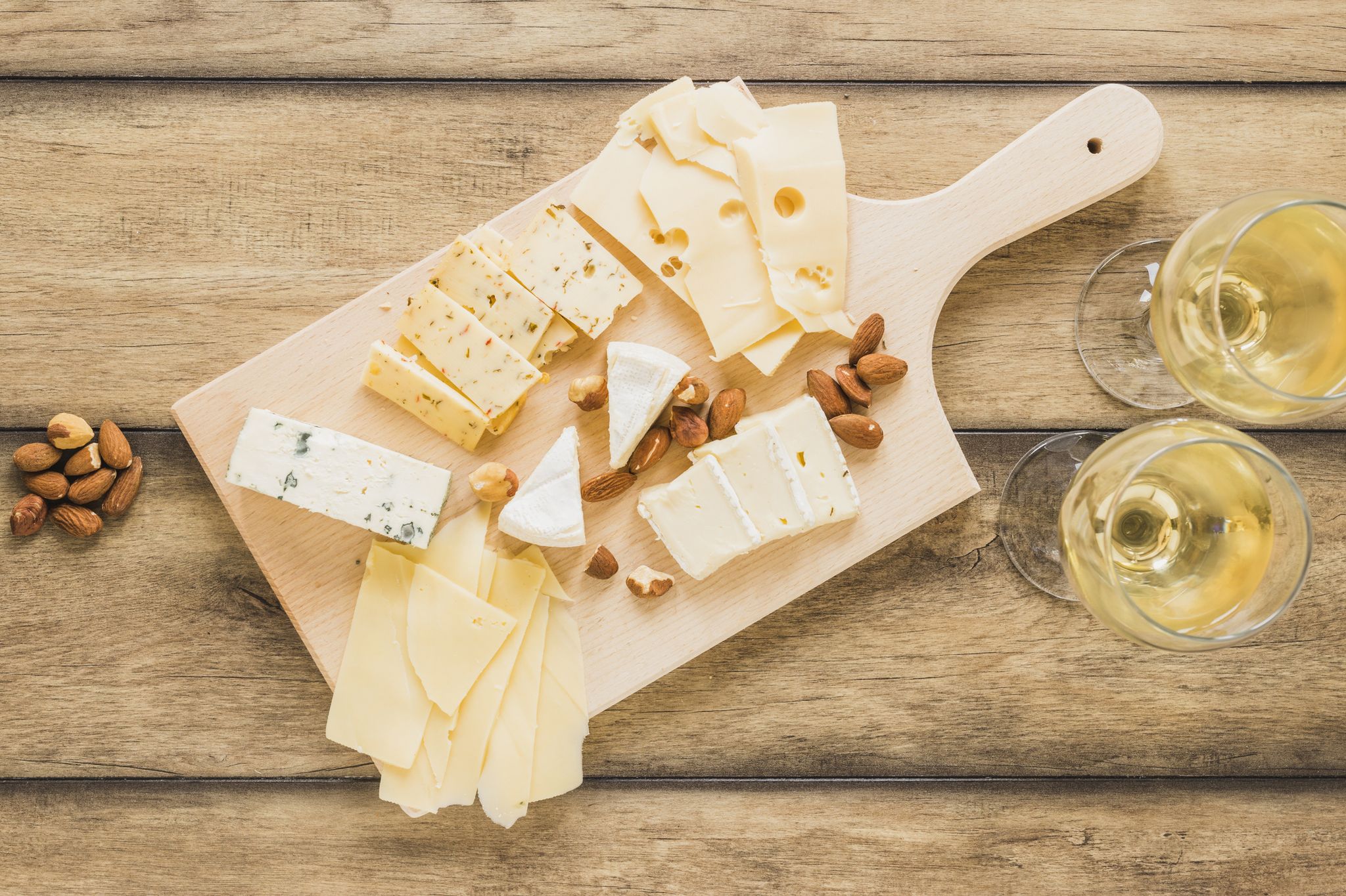 cheese board with various cheeses placed on wooden counter top with wine and bowl of nuts