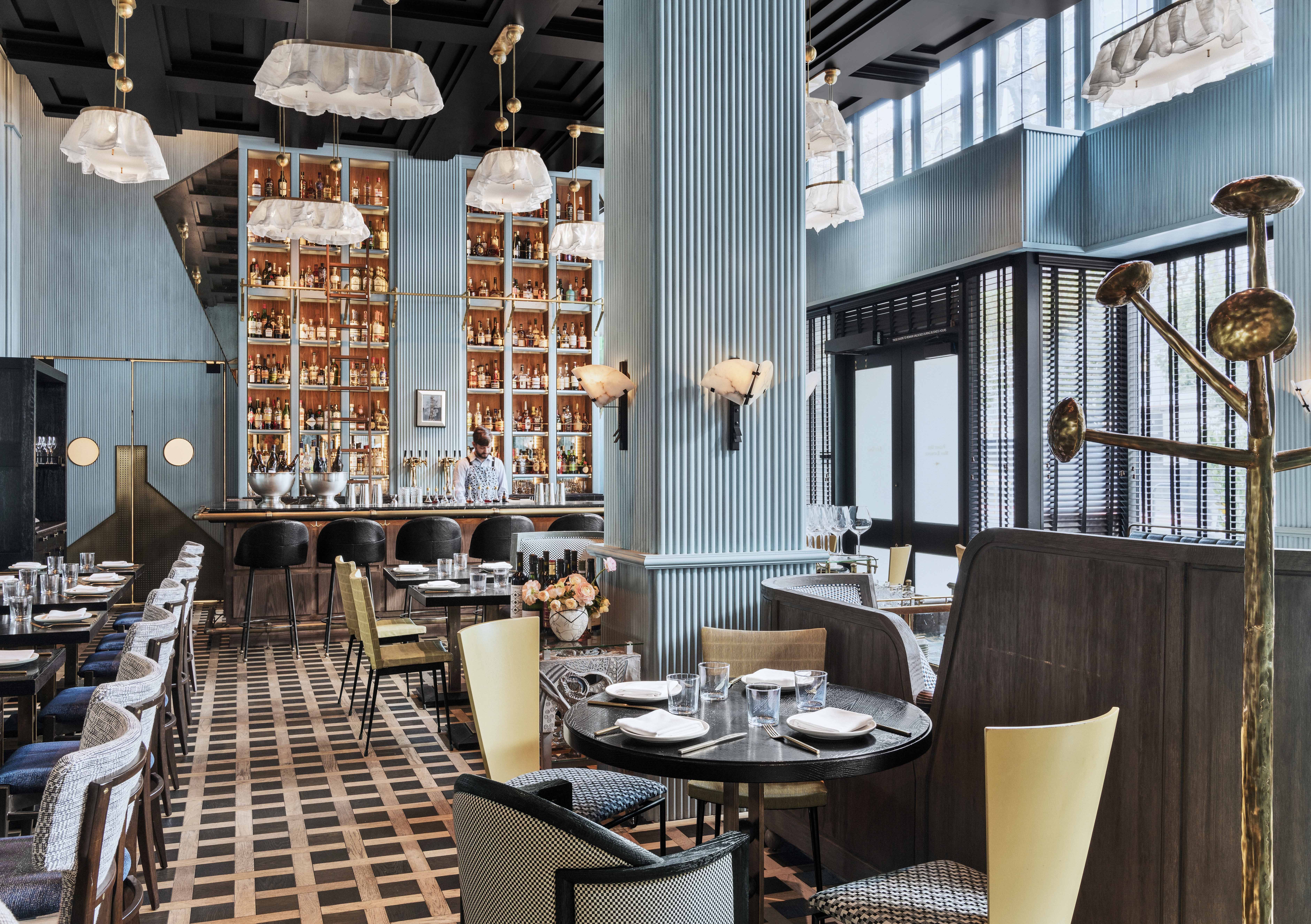 A List 2022 - ELLE DECOR restaurant interior with wood floors table banquettes blue pillars and a large wall of bottles and glass at the far end behind the bar