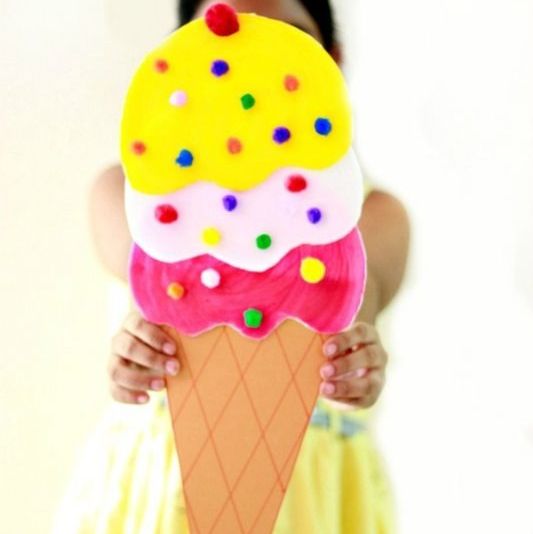 a child holds up a giant ice cream cone craft, with three scoops and colored pom poms as sprinkles
