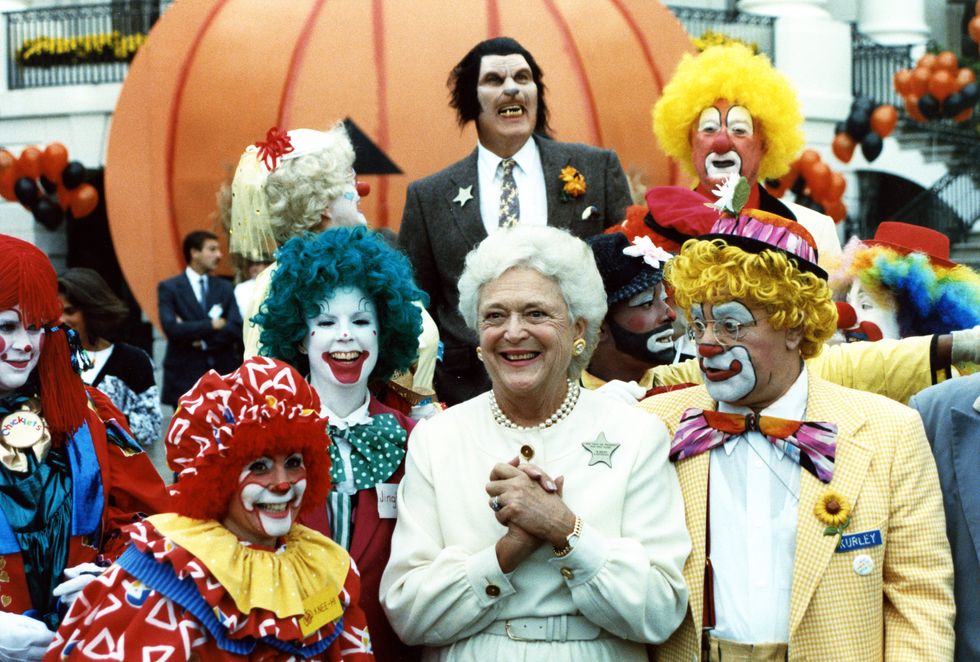 this photograph is of first lady barbara bush grinning as she poses with costumed performers, a group of clowns, and a lone werewolf on the south grounds of the white house as part of a halloween celebration