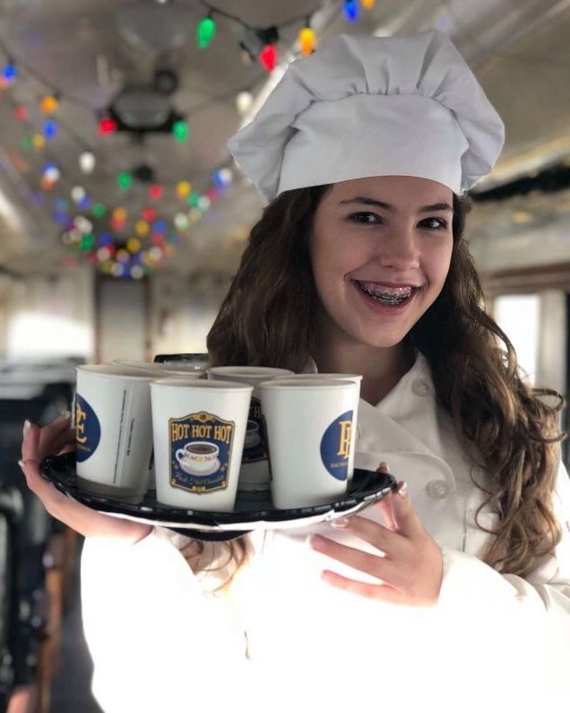 young girl in a chef uniform and hat smiling widely as she holds up a silver tray with cups of hot chocolate that have a picture of hot chocolate and say "hot hot hot" on them