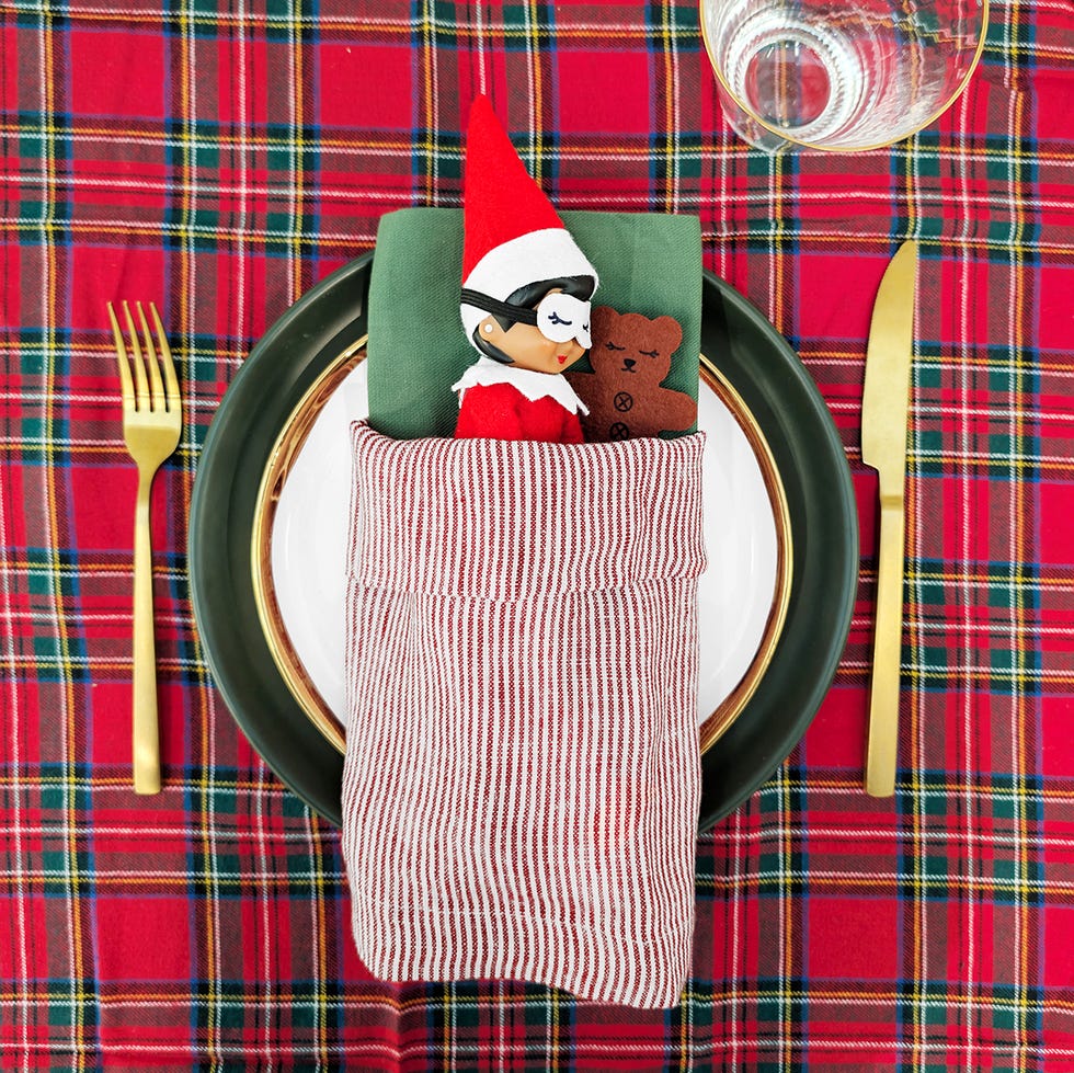 an elf sleeps in the middle of a table setting, using a napkin as a sleeping bag and wearing a teeny eye mask