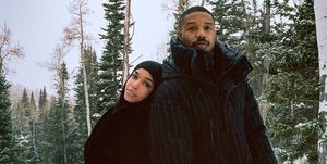 michael b jordan and lori harvey dressed in black coats, leaning on each other in front of green pine trees in the snow