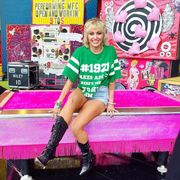 miley cyrus before the super bowl, wearing a blonde mullet and green t shirt, sitting on a barbie pink pool table with collages in the background