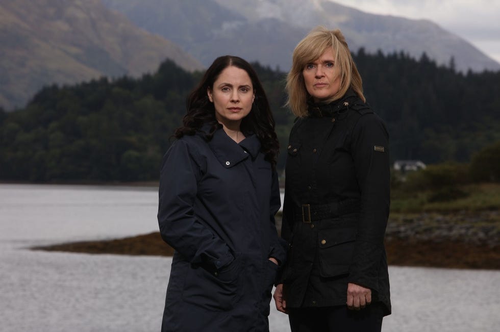 two women standing by a body of water with a mountainous backdrop